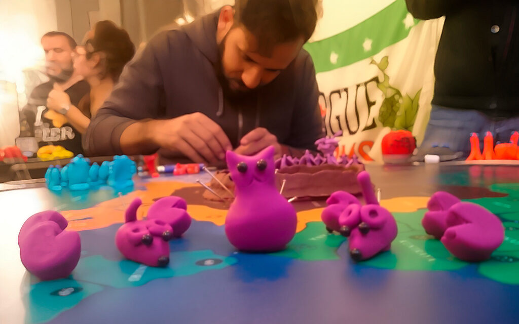 Criaturas de massinha roxa recém-criadas sobre o tabuleiro do Planeta Insano, enquanto um jogador molda novas formas ao fundo durante a partida.