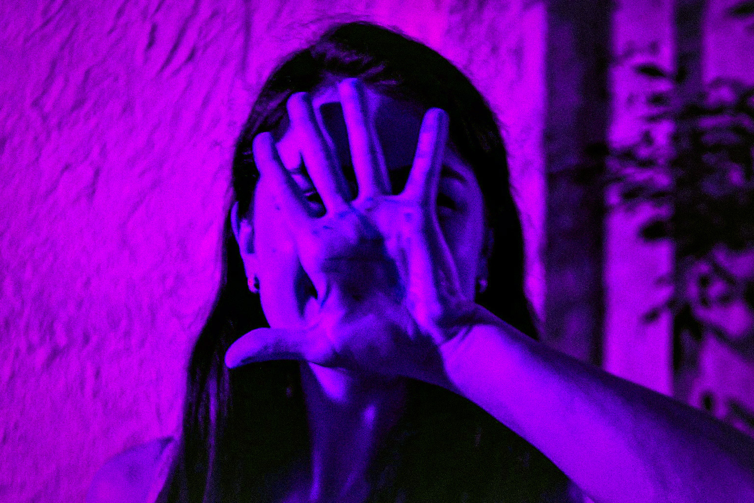 Pessoa com a mão estendida à frente do rosto, parcialmente ocultando a expressão, em gesto teatral iluminado por luz quente, evocando mistério e presença artística.