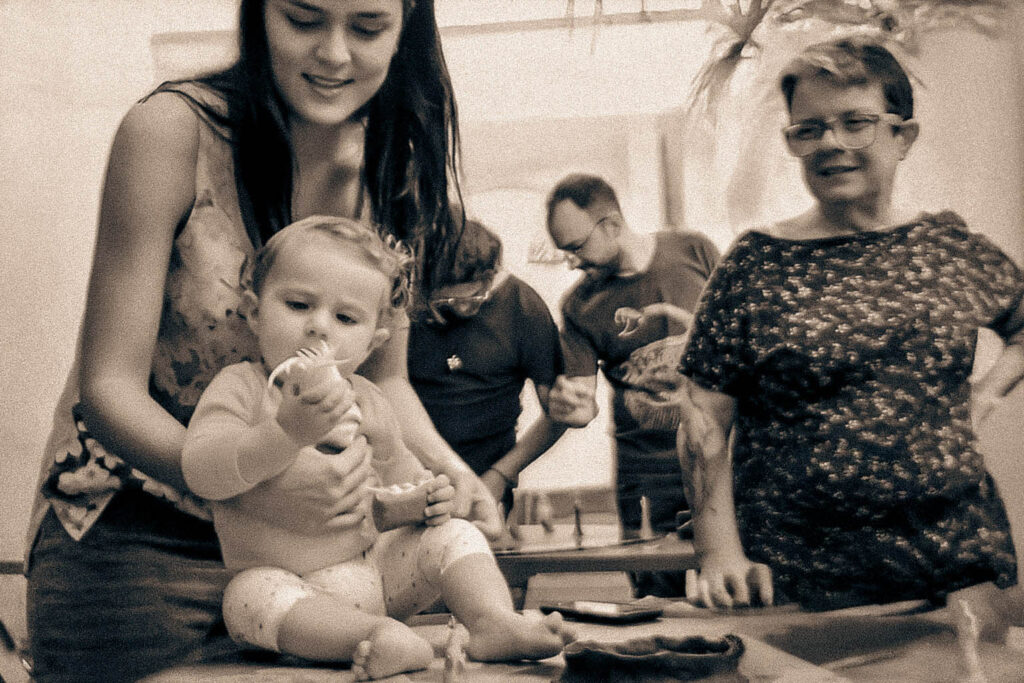 Bebê sentado sobre uma mesa, segurando um objeto com curiosidade, amparado por uma pessoa adulta, enquanto outras pessoas observam ao fundo em um ambiente artístico coletivo.