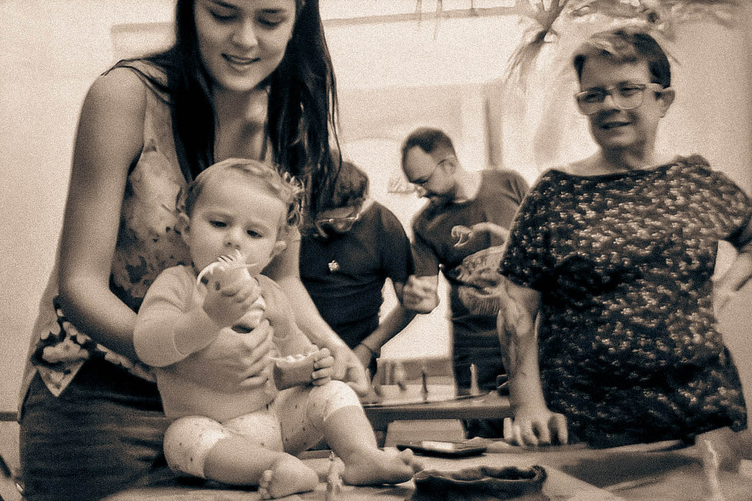 Bebê sentado sobre uma mesa, segurando um objeto com curiosidade, amparado por uma pessoa adulta, enquanto outras pessoas observam ao fundo em um ambiente artístico coletivo.