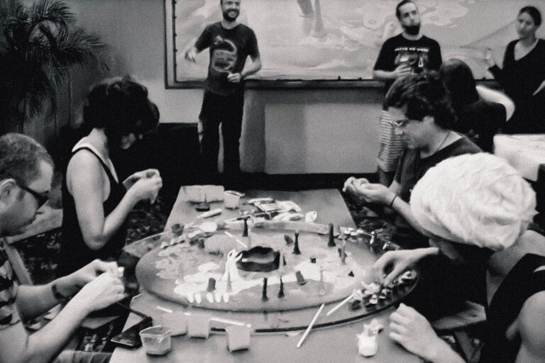 Pessoas reunidas ao redor da mesa circular do Planta Insano, moldando peças e elementos artísticos coletivamente, enquanto outras observam e interagem ao fundo em um espaço cultural.