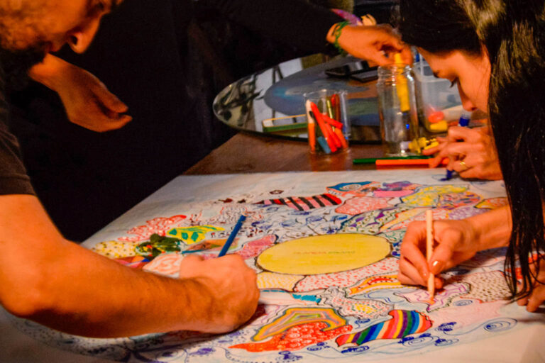 Duas pessoas desenhando juntas sobre uma grande ilustração circular colorida, com lápis e canetas marcando caminhos diferentes no mesmo mapa artístico.