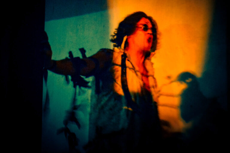 Pessoa em performance teatral com iluminação dramática em tons quentes e frios, corpo em movimento e expressão intensa, evocando presença ritual e força simbólica.