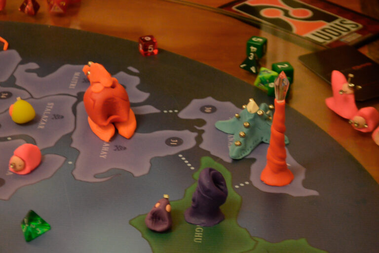 Criaturas coloridas do jogo Planeta Insano espalhadas pelo tabuleiro durante partida na Ludos Luderia, com dados e torres energéticas ao fundo.