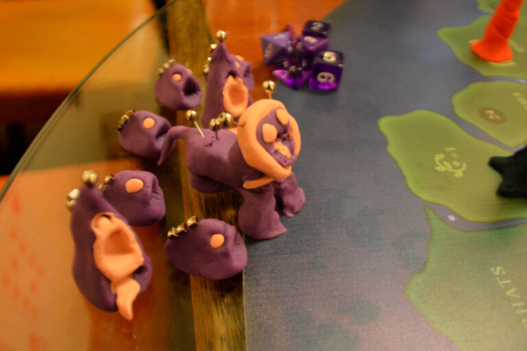 Criaturas roxas do Planeta Insano reunidas à beira do tabuleiro durante partida na Ludos Luderia, observando o território enquanto dados de RPG aguardam o próximo movimento.