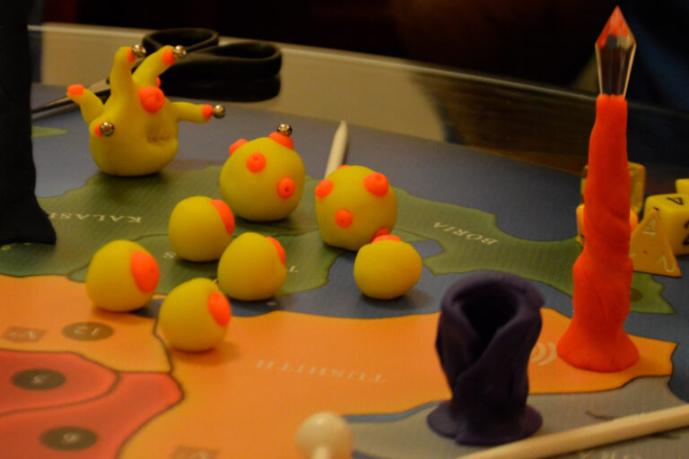 Criaturas amarelas do Planeta Insano reunidas próximas a um Altar da Transmutação durante partida do jogo de tabuleiro na Ludos Luderia.