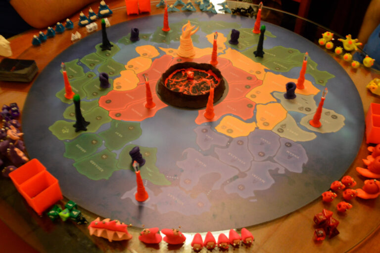 Tabuleiro do jogo de tabuleiro Planeta Insano completamente montado na Ludos Luderia, com espécies dos jogadores posicionadas na borda, nove altares da transmutação, criatura ancestral no deserto, árvores da vida eterna e buracos de minhoca espalhados pelo mapa.