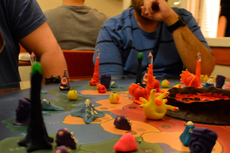 Jogadores observam o tabuleiro do jogo Planeta Insano durante a fase inicial, com criaturas coloridas distribuídas pelo mapa, altares da transmutação ativos e o núcleo vulcânico ao centro.