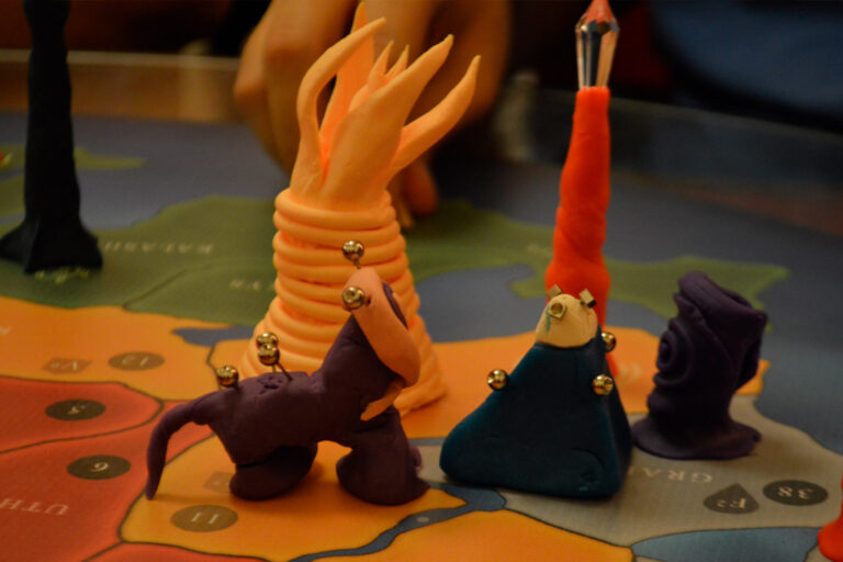 Criaturas do jogo Planeta Insano reunidas próximas à criatura ancestral e a um altar da transmutação, durante partida na Ludos Luderia, representando um momento ritualístico do tabuleiro.