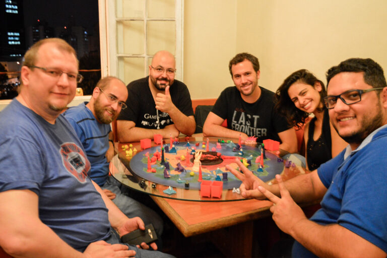 Grupo de jogadores reunidos ao redor do tabuleiro do jogo Planeta Insano na Ludos Luderia, com criaturas, altares da transmutação e o planeta completamente montado durante uma partida coletiva.