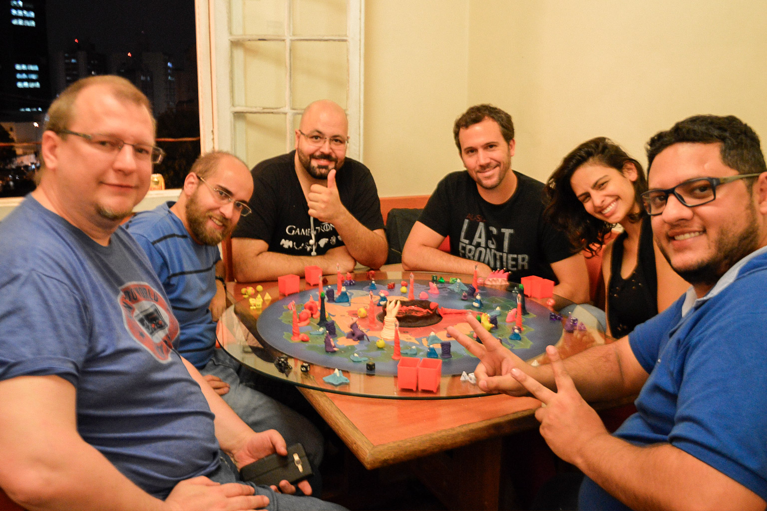 Grupo de jogadores reunidos ao redor do tabuleiro do jogo Planeta Insano na Ludos Luderia, com criaturas, altares da transmutação e o planeta completamente montado durante uma partida coletiva.