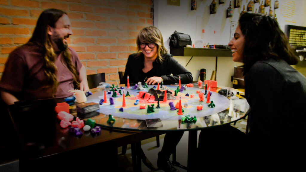 Grupo de amigos jogando e se divertindo em torno de um jogo de tabuleiro criativo, com miniaturas coloridas e clima descontraído e artístico.