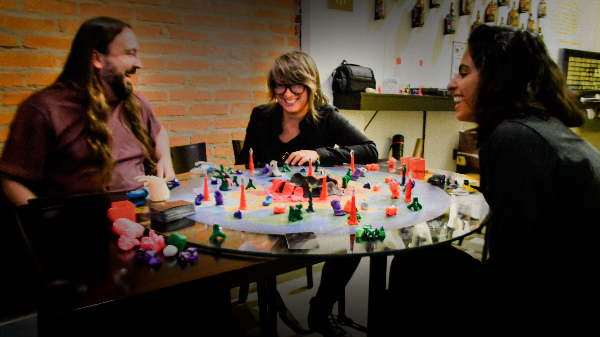 Grupo de amigos jogando e se divertindo em torno de um jogo de tabuleiro criativo, com miniaturas coloridas e clima descontraído e artístico.