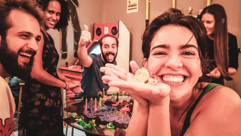 Grupo de amigos jogando e se divertindo em torno de um jogo de tabuleiro criativo, com miniaturas coloridas e clima descontraído e artístico.