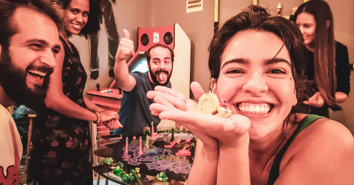 Grupo de amigos jogando e se divertindo em torno de um jogo de tabuleiro criativo, com miniaturas coloridas e clima descontraído e artístico.
